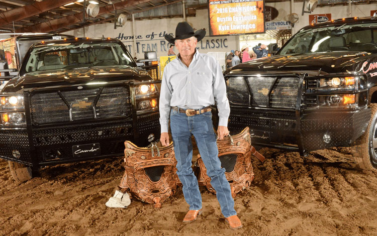 George Strait standing in front of two Chevy trucks and posing with the winners saddles for the George Strait Team Roping Classic.